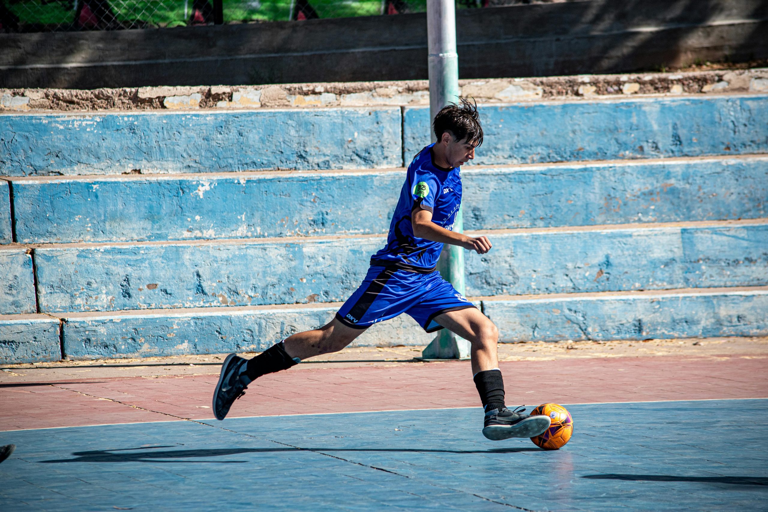 Teenage boy in blue jersey playing soccer on an outdoor court, mid-action.